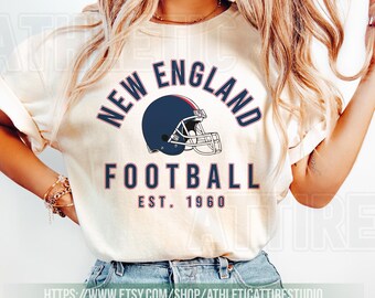 May include: Ivory-colored t-shirt featuring the text "NEW ENGLAND FOOTBALL" above a navy blue football helmet graphic. The text "EST. 1960" is below the graphic. The shirt is worn with blue jeans.