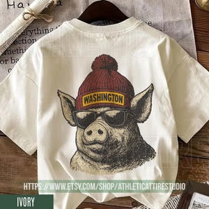 May include: Ivory-colored t-shirt featuring a graphic of a pig wearing a maroon beanie with the word "WASHINGTON" on it, and sunglasses. The pig graphic is in black and white. The t-shirt has a crew neck and short sleeves.