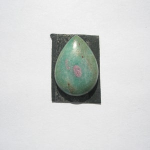 May include: A teardrop-shaped green gemstone with pink and gold inclusions. The gemstone is set in a black backing.