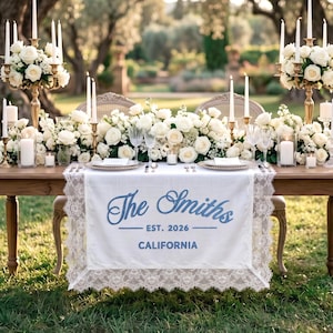 May include: A beautifully decorated wedding table with white roses, candles, and gold candelabras. The table has a white runner with the text "The Smiths EST. 2026 CALIFORNIA" in blue script. The table is set for two, creating a romantic ambiance.