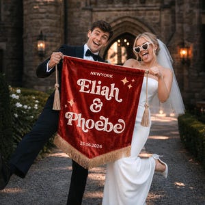 May include: A deep red velvet pennant banner featuring gold embroidered text that reads NEWYORK, Eliah & Phoebe, and 25.06.2026. The banner is decorated with gold star accents, gold tassels on the top corners, and gold fringe along the bottom edge.