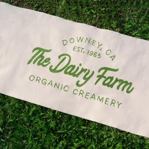 Könnte beinhalten: Weiße Stofftasche mit grüner Stickerei, auf der "The Dairy Farm, Organic Creamery, Downey, CA, Est. 1965" steht.