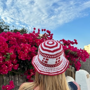 May include: A red and white crocheted bucket hat with a circular pattern. The hat is worn by a person with long blonde hair, standing in front of a bush of pink flowers.