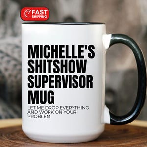 May include: White ceramic mug with a black handle and rim. The mug features the text "MICHELLE'S SHITSHOW SUPERVISOR MUG" in bold black letters, with the phrase "LET ME DROP EVERYTHING AND WORK ON YOUR PROBLEM" below. A red "FAST SHIPPING" sticker is visible.