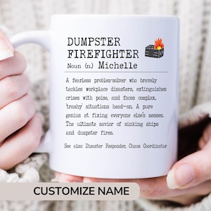 May include: White ceramic mug with a black and white definition of "Dumpster Firefighter" with a small illustration of a burning dumpster. The definition reads: "A fearless problem-solver who bravely tackles workplace disasters, extinguishes crises with poise, and faces complex, 'trashy' situations head-on. A pure genius at fixing everyone else's messes. The ultimate savior of sinking ships and dumpster fires. See also: Disaster Responder, Chaos Coordinator." The mug has the name "Michelle" in the definition. The text "CUSTOMIZE NAME" is at the bottom of the mug.