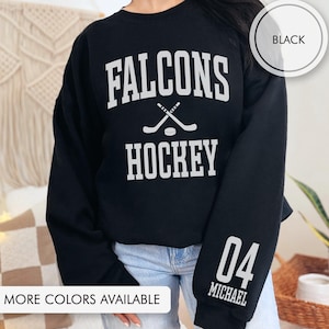 May include: Black sweatshirt with white text reading "Falcons Hockey" with crossed hockey sticks and a puck. The sleeve has the number "04" and the name "Michael". The words "More Colors Available" are at the bottom.
