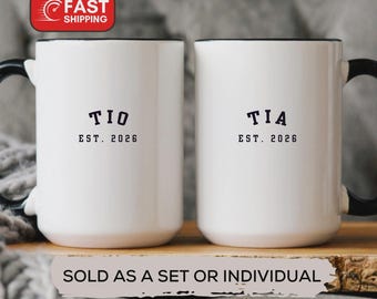 May include: Two white ceramic mugs with black handles and rims. One mug reads "TIO EST. 2026" and the other reads "TIA EST. 2026" in black text. The mugs are displayed on a wooden surface.