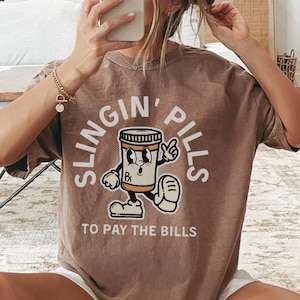 May include: A brown t-shirt with the words "SLINGIN' PILLS" and "TO PAY THE BILLS" printed on it. The graphic features a cartoon pill bottle character. The shirt is short-sleeved and has a relaxed fit.