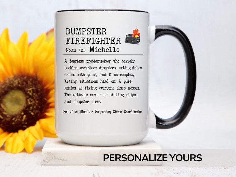 May include: White ceramic mug with a black handle. The mug has a black and white design that reads "DUMPSTER FIREFIGHTER Noun (n.) Michelle A fearless problem-solver who bravely tackles workplace disasters, extinguishes crises with poise, and faces complex, trashy situations head-on. A pure genius at fixing everyone else's messes. The ultimate savior of sinking ships and dumpster fires. See also Disaster Responder, Chaos Coordinator"