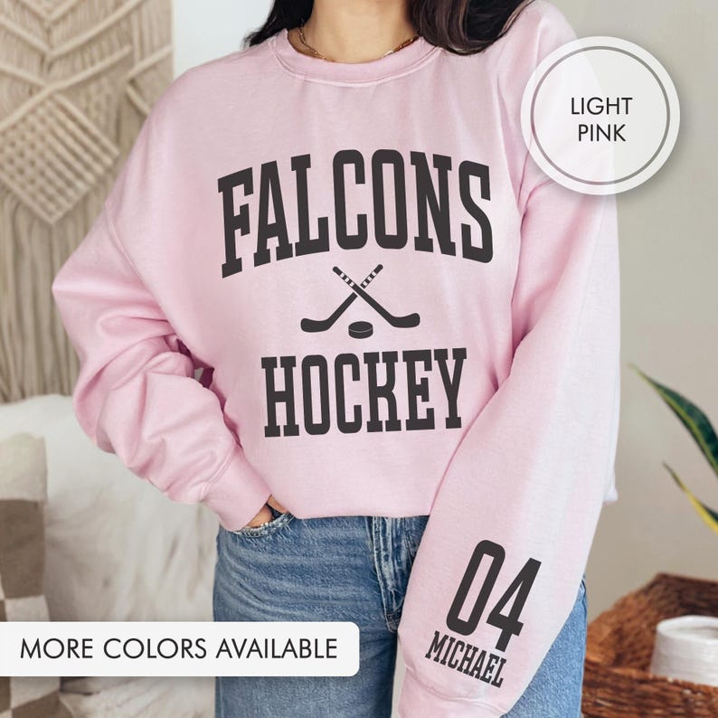 May include: Pale pink sweatshirt with the text "FALCONS HOCKEY" in black. The design includes crossed hockey sticks and a puck. The sleeve features the number "04" and the name "MICHAEL". The text "MORE COLORS AVAILABLE" is at the bottom.