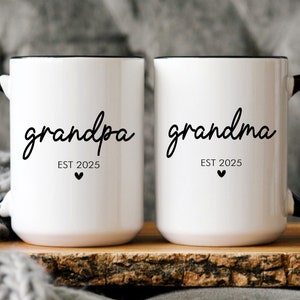 May include: Two white ceramic mugs with black handles and black rims. The mug on the left says "grandpa EST 2025" with a small heart below the text. The mug on the right says "grandma EST 2025" with a small heart below the text.