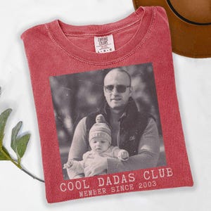 K&ouml;nnte beinhalten: Rotes Comfort Colors T-Shirt mit einem Schwarz-Wei&szlig;-Foto eines Mannes und eines Babys. Der Text "COOL DADAS CLUB MEMBER SINCE 2003" ist unter dem Foto aufgedruckt. Ein brauner Hut ist oben rechts zu sehen.