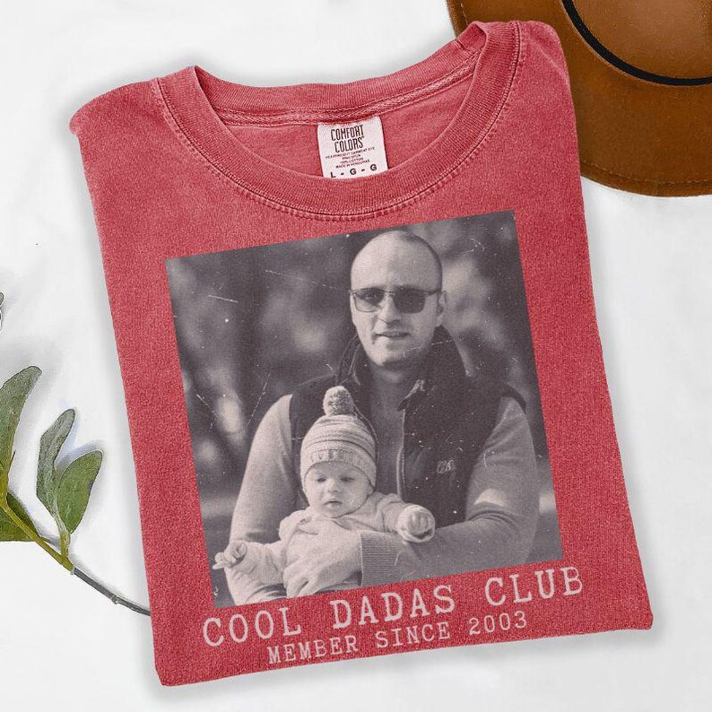K&ouml;nnte beinhalten: Rotes Comfort Colors T-Shirt mit einem Schwarz-Wei&szlig;-Foto eines Mannes und eines Babys. Der Text "COOL DADAS CLUB MEMBER SINCE 2003" ist unter dem Foto aufgedruckt. Ein brauner Hut ist oben rechts zu sehen.