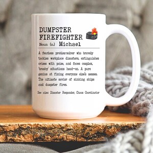 May include: White ceramic mug with a black and white design that reads "DUMPSTER FIREFIGHTER Noun (n.) Michael A fearless problem-solver who bravely tackles workplace disasters, extinguishes crises with poise, and faces complex, trashy situations head-on. A pure genius at fixing everyone else's messes. The ultimate savior of sinking ships and dumpster fires. See also Disaster Responder, Chaos Coordinator"