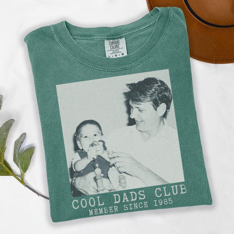 K&ouml;nnte beinhalten: Gr&uuml;nes Comfort Colors T-Shirt mit einem Schwarz-Wei&szlig;-Foto. Das Foto zeigt eine Person, die ein Baby h&auml;lt. Der Text "COOL DADS CLUB MEMBER SINCE 1985" ist darunter gedruckt.