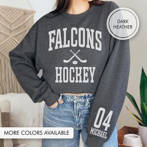 May include: Dark grey sweatshirt with the text "Falcons Hockey" in white, featuring crossed hockey sticks and a puck. The right sleeve displays the number "04" and the name "Michael" in white. The phrase "More Colors Available" is at the bottom of the image.