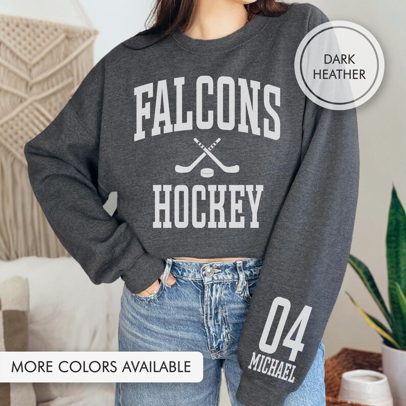 May include: Dark grey sweatshirt with the text "Falcons Hockey" in white, featuring crossed hockey sticks and a puck. The right sleeve displays the number "04" and the name "Michael" in white. The phrase "More Colors Available" is at the bottom of the image.