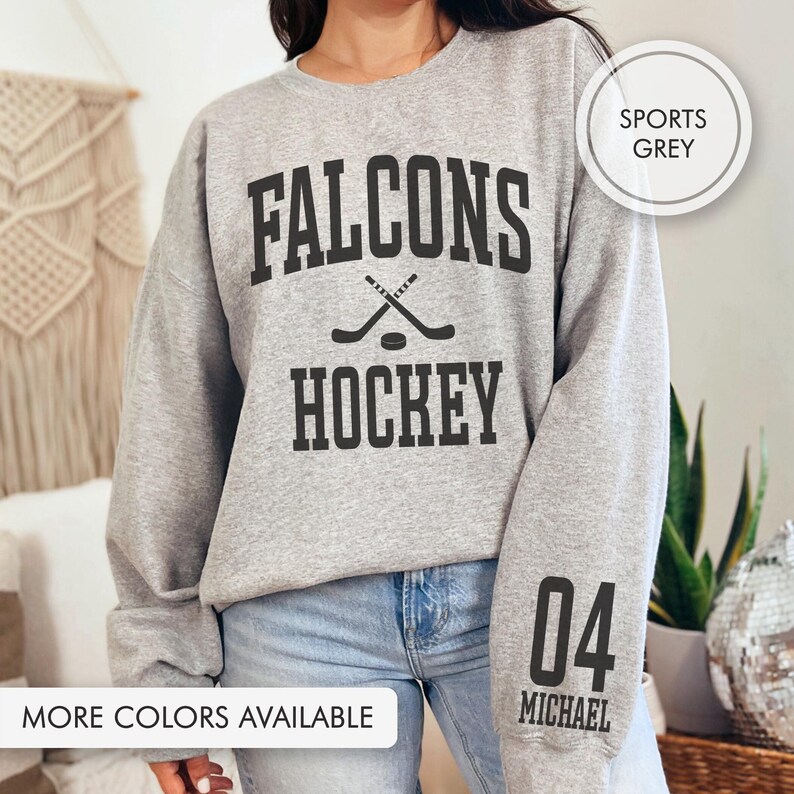 May include: A light grey sweatshirt featuring the text "FALCONS HOCKEY" in black, with crossed hockey sticks. The sleeve displays the number "04" and the name "MICHAEL". The phrase "MORE COLORS AVAILABLE" is visible at the bottom.