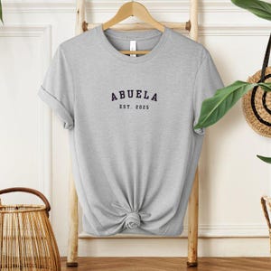 May include: A gray t-shirt with the text "Abuela Est. 2025" printed on the front.