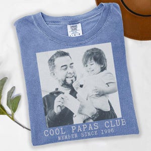 K&ouml;nnte beinhalten: Ein blaues Comfort Colors T-Shirt mit einem Schwarz-Wei&szlig;-Bild von Vater und Kind. Der Text "COOL PAPAS CLUB MEMBER SINCE 1996" ist darunter gedruckt. Ein brauner Hut ist in der oberen rechten Ecke zu sehen.
