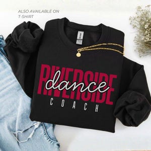 May include: Black sweatshirt with the text "Riverside Dance Coach" in a red and white design. A gold necklace with a pendant is draped over the sweatshirt. The words "Also available on T-shirt" are in the upper left corner.