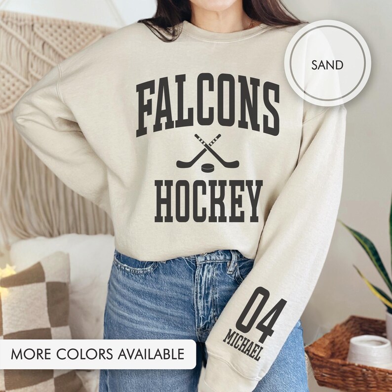 May include: A sand-coloured sweatshirt featuring the text "FALCONS HOCKEY" in black, with hockey sticks and a puck graphic. The right sleeve has "04 MICHAEL" in black. The sweatshirt is worn with blue jeans.