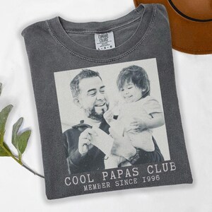K&ouml;nnte beinhalten: Dunkelgraues T-Shirt mit einem Schwarz-Wei&szlig;-Foto eines Mannes und eines Kindes. Der Text "COOL PAPAS CLUB MEMBER SINCE 1996" ist unten aufgedruckt. Das T-Shirt ist auf einer wei&szlig;en Oberfl&auml;che gefaltet.