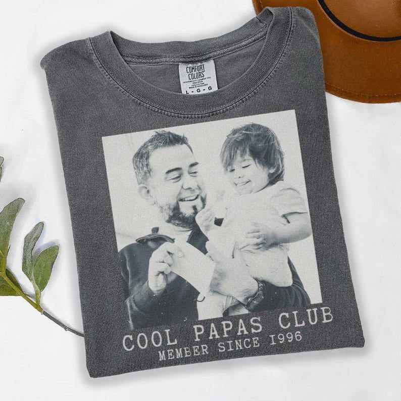 K&ouml;nnte beinhalten: Dunkelgraues T-Shirt mit einem Schwarz-Wei&szlig;-Foto eines Mannes und eines Kindes. Der Text "COOL PAPAS CLUB MEMBER SINCE 1996" ist unten aufgedruckt. Das T-Shirt ist auf einer wei&szlig;en Oberfl&auml;che gefaltet.