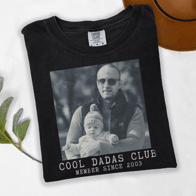 K&ouml;nnte beinhalten: Schwarzes T-Shirt mit einem Schwarz-Wei&szlig;-Foto eines Mannes, der ein Baby h&auml;lt. Der Text "COOL DADAS CLUB MEMBER SINCE 2003" ist unter dem Foto aufgedruckt. Das Shirt besteht aus einem weichen Material.