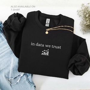 May include: Black crewneck sweatshirt with the text "in data we trust" in white, along with a graphic of a bar chart. A gold necklace with a round pendant rests on the sweatshirt. The words "ALSO AVAILABLE ON T-SHIRT" are visible.