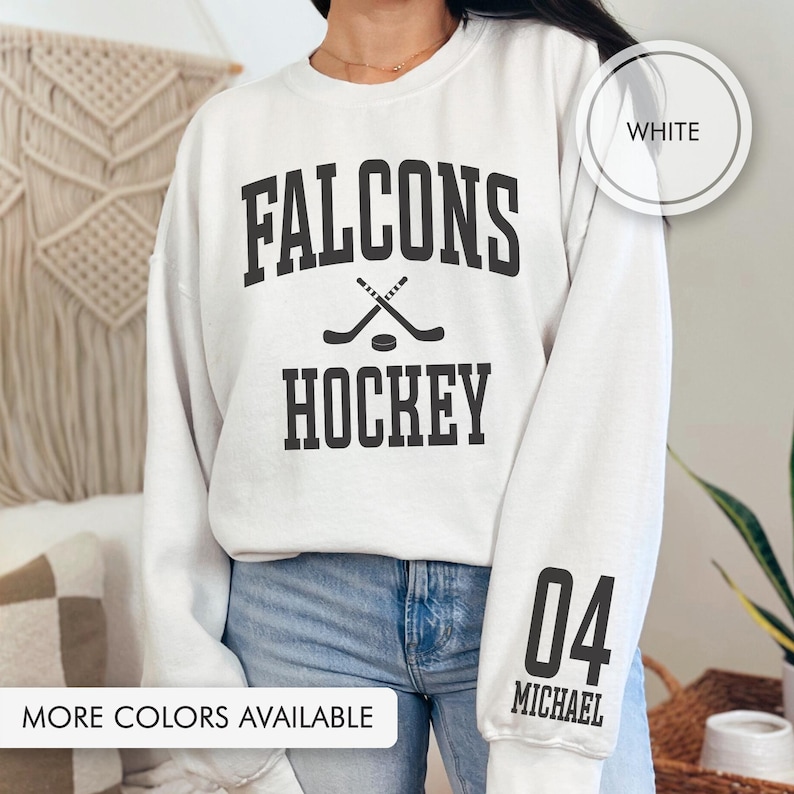 May include: Cream-coloured sweatshirt featuring the text "FALCONS HOCKEY" in black, with crossed hockey sticks and a puck graphic. The right sleeve has "04 MICHAEL" in black. The text "MORE COLORS AVAILABLE" is at the bottom. A white circle with "WHITE" is in the upper right.