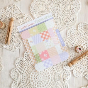 May include: A craft-themed image featuring a "Pastel Prairie Binding Holder" with colorful patterned squares. Wooden spools and clothespin-style clips are arranged on white lace doilies, creating a vintage aesthetic.