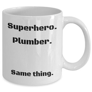 Puede incluir: Taza de cerámica blanca con asa curva. La taza presenta el texto "Superhero. Plumber. Same thing." en letras negras en negrita. Diseñada para bebidas calientes.