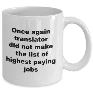 May include: White ceramic coffee mug with a curved handle. The mug features black text that reads: "Once again translator did not make the list of highest paying jobs."