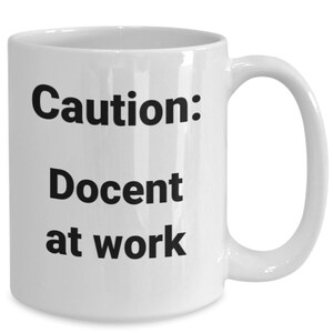 May include: White ceramic coffee mug with black text that reads "Caution: Docent at work".