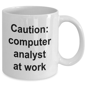 Może przedstawiać: Biały ceramiczny kubek do kawy z czarnym tekstem, który brzmi "Uwaga: analityk komputerowy w pracy".