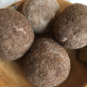May include: Four felted wool balls in a wooden bowl. Two of the balls are brown and two are a light brown color.