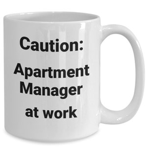 May include: White ceramic coffee mug with black text that reads "Caution: Apartment Manager at work".