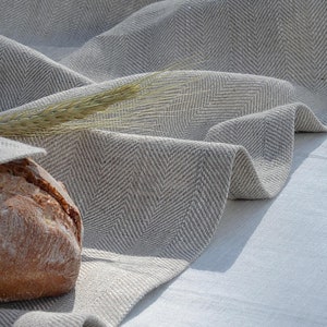 Op de afbeelding: Een lichtgrijze visgraat linnen tafelkleed met een brood en tarwearen. De stof drapeert over een wit oppervlak en creëert zachte plooien en schaduwen. Het brood heeft een goudbruine korst.