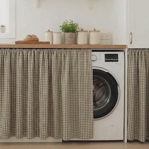 May include: A white washing machine is partially visible behind a tan and white checkered curtain. The curtain hangs below a wooden countertop. A loaf of bread, potted plant, and canisters sit on the countertop.