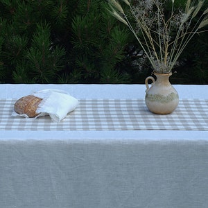 Könnte beinhalten: Eine weiße Leinen-Tischdecke mit einem beige-weißen karierten Läufer. Ein Brotlaib ist in ein weißes Tuch auf dem Läufer gewickelt. Eine braune Keramikvase mit getrockneten Blumen steht auf dem Läufer.