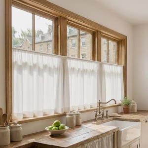 Puede incluir: Una ventana de cocina con marco de madera y cortinas blancas. La ventana da a un edificio. Una encimera de madera sostiene un fregadero blanco, un cuenco de manzanas verdes y tarros de cerámica.