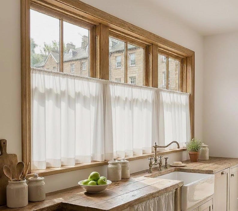 May include: A kitchen window with a wooden frame and white curtains. The window overlooks a building. A wooden countertop holds a white sink, a bowl of green apples, and ceramic jars.