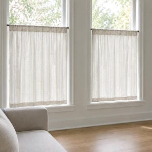 May include: Two cafe-style window curtains featuring a light beige and white vertical pinstripe pattern. Each curtain is hung on a thin, dark-toned rod mounted inside the window frame, covering the lower half of the window glass.