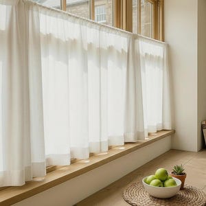 May include: Sheer white curtains hang in front of a window, filtering sunlight. A wooden window sill holds a bowl of green apples and a small potted plant. The room has a bright, airy feel.