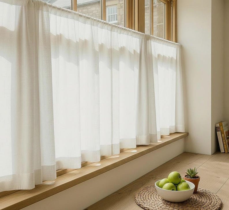 May include: Sheer white curtains hang in front of a window, filtering sunlight. A wooden window sill holds a bowl of green apples and a small potted plant. The room has a bright, airy feel.