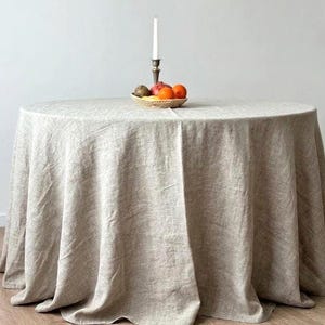 Op de afbeelding: Een ronde, beige linnen tafelkleed bedekt een houten tafel. Een klein gevlochten mandje bevat fruit en een kaars staat op een decoratieve kandelaar. De tafel staat tegen een effen witte muur.
