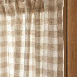 May include: A semi-sheer curtain panel featuring a light beige and white checked pattern. The fabric has a visible woven texture and hangs from a simple rod, creating soft vertical folds that allow light to pass through the material.