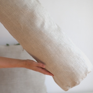 May include: A large, beige, cylindrical pillow with a herringbone pattern. The pillow is being held by a hand.
