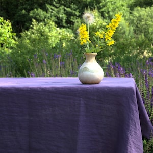 Pode incluir: Um pequeno vaso de cerâmica branco-sujo com flores amarelas e uma toalha de mesa roxa. O vaso é redondo com uma superfície texturizada. A toalha de mesa é de cor roxa escura lisa. O fundo apresenta folhagem verde e flores roxas.